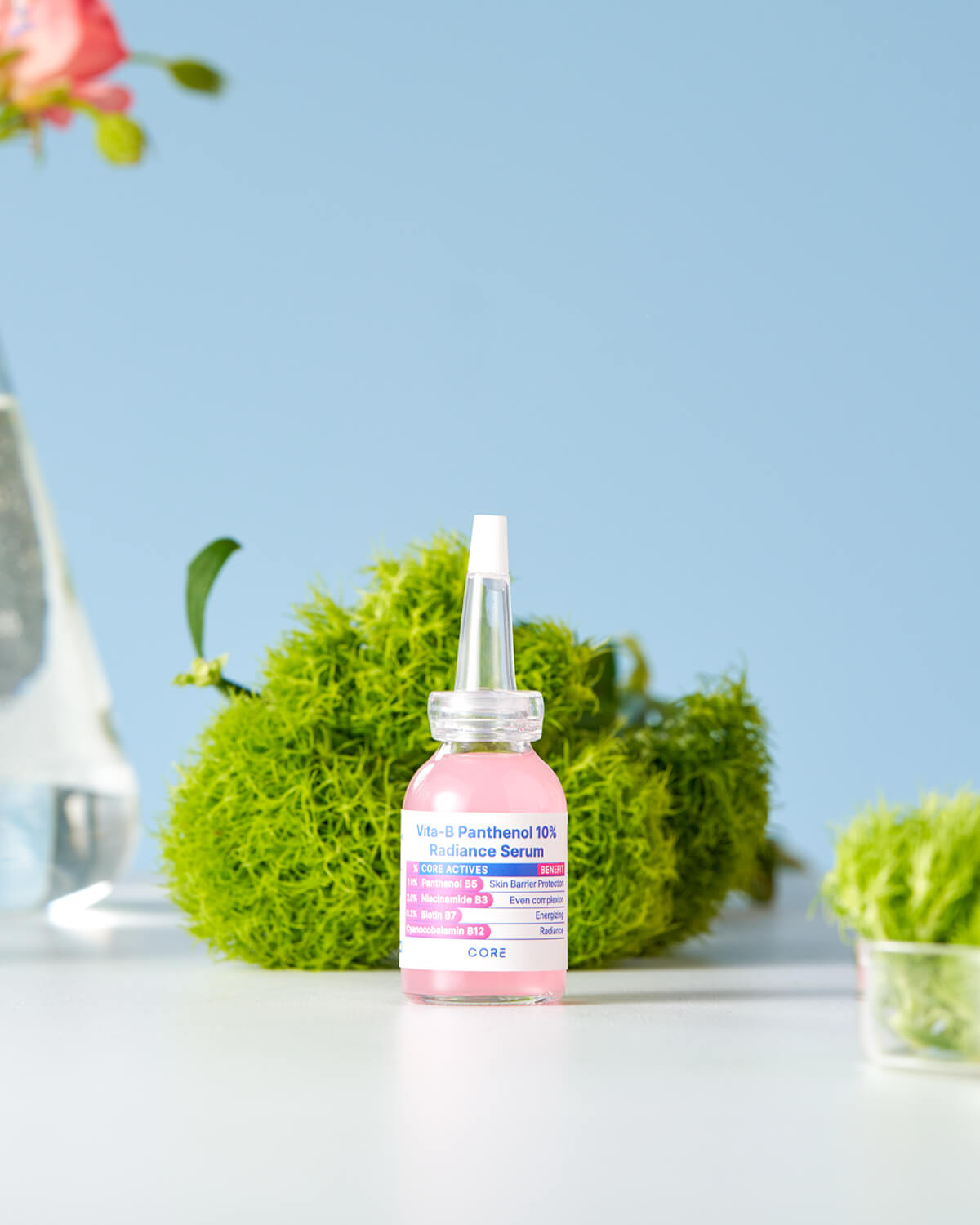 The serum bottle is featured in a still life, standing on a white surface next to a bright green, moss-like object and a clear glass flask, with a pink flower element in the upper corner.