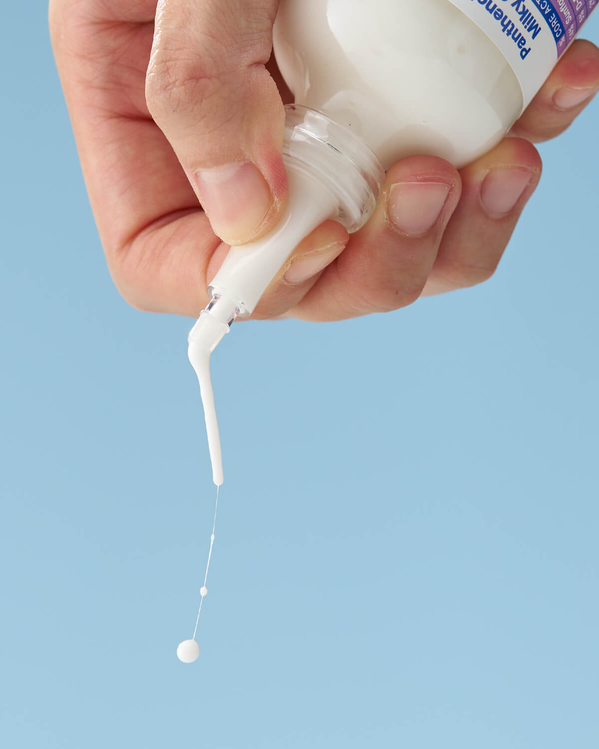 A hand holds the product bottle, allowing a thin stream of the white cream to drip from the nozzle tip, creating small, hanging drops against a blue background.