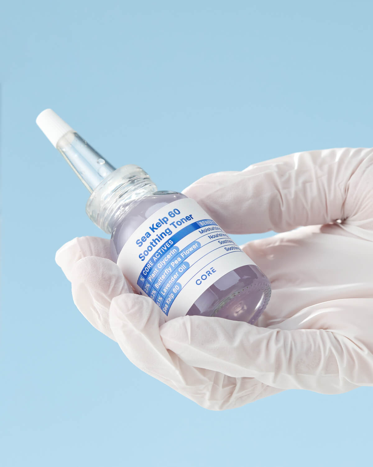 A hand wearing a white medical glove holds the small, clear bottle of Core Sea Kelp 60 Soothing Toner against a light blue background, showcasing the product in a clinical setting.