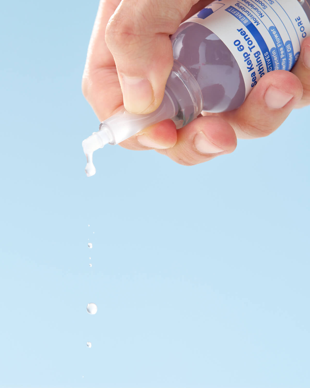A hand holds the toner bottle upside down, with a few drops of the pale liquid dripping from the nozzle tip onto a light blue background.