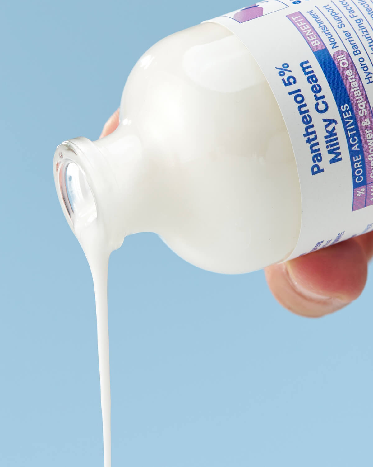 A close-up shot of the white, milky cream product being poured out of the bottle's wide mouth, creating a thick stream against a light blue background.