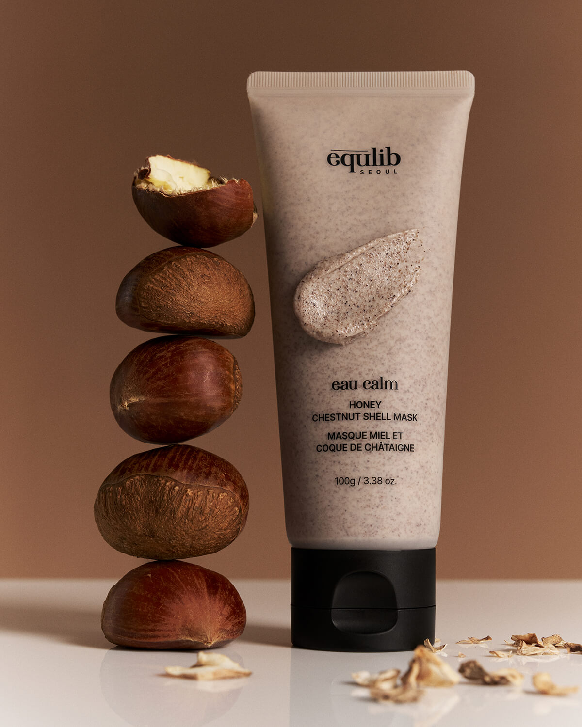 A tube of Equlib Eau Calm Honey Chestnut Shell Mask standing next to a vertical stack of whole raw chestnuts.