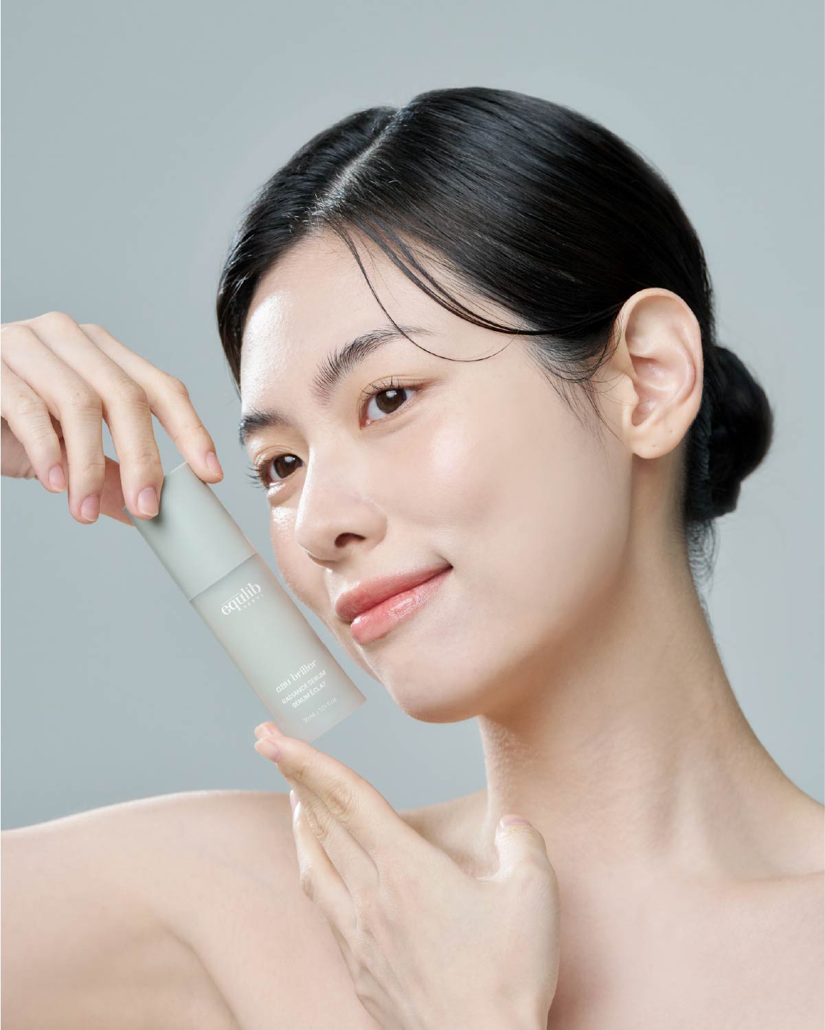 A smiling East Asian woman with clear, glowing skin holding the Equlib radiance serum bottle next to her cheek.