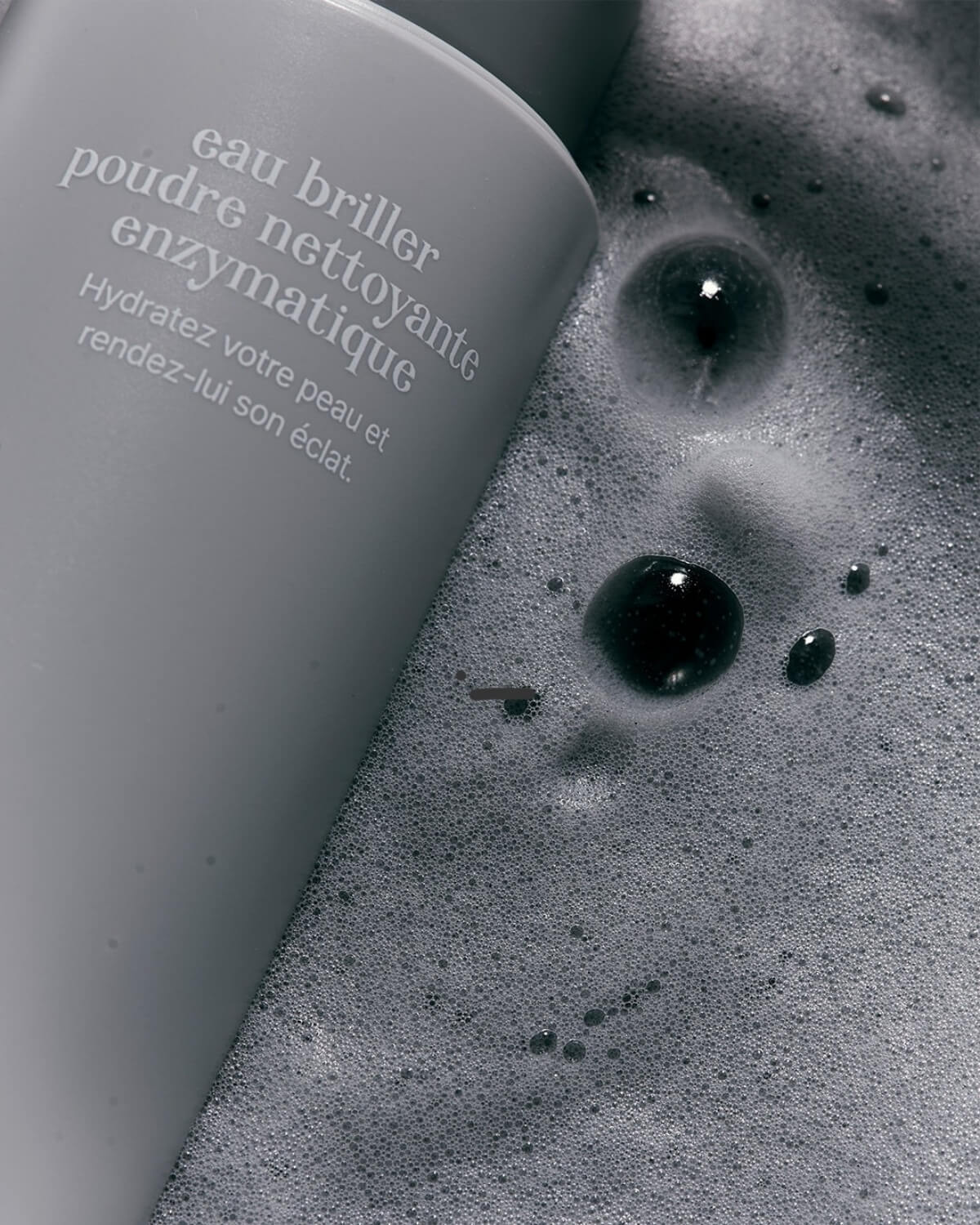 Overhead macro shot of Equlib skincare cleanser bottle immersed in bubbly, dark grey charcoal foaming lather.