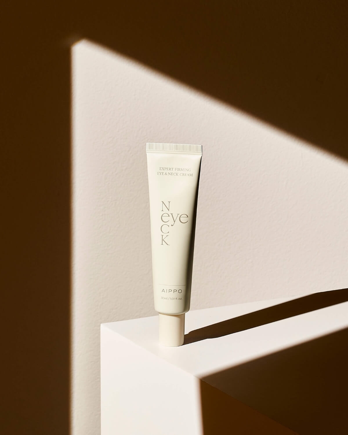 A single tube of the Eye & Neck Cream stands on a white geometric platform, dramatically lit by a diagonal beam of sunlight that casts a sharp shadow.