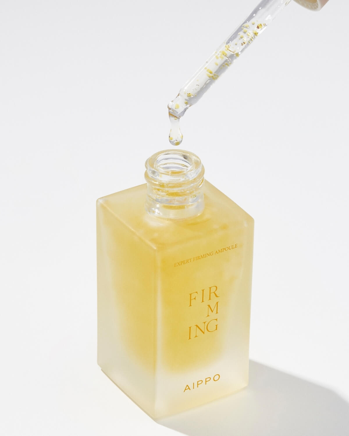 A close-up shot of the AIPPO Expert Firming Ampoule bottle with the dropper being pulled out. A drop of the yellow, bubbly product is visibly dripping from the pipette back into the bottle's neck. The label "EXPERT FIRMING AMPOULE" is visible on the side.