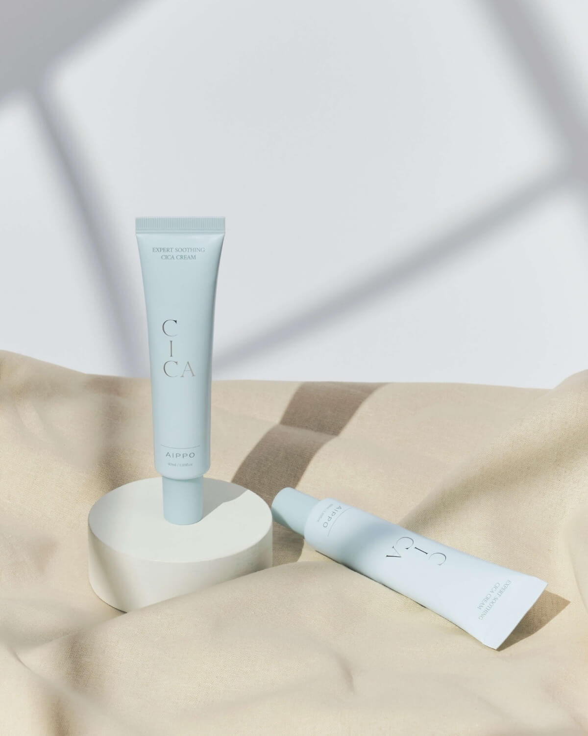 Two tubes of the Expert Soothing Cica Cream are displayed: one standing upright on a white cylindrical pedestal and the other lying flat on a soft, draped beige fabric, lit by a strong, directional light.