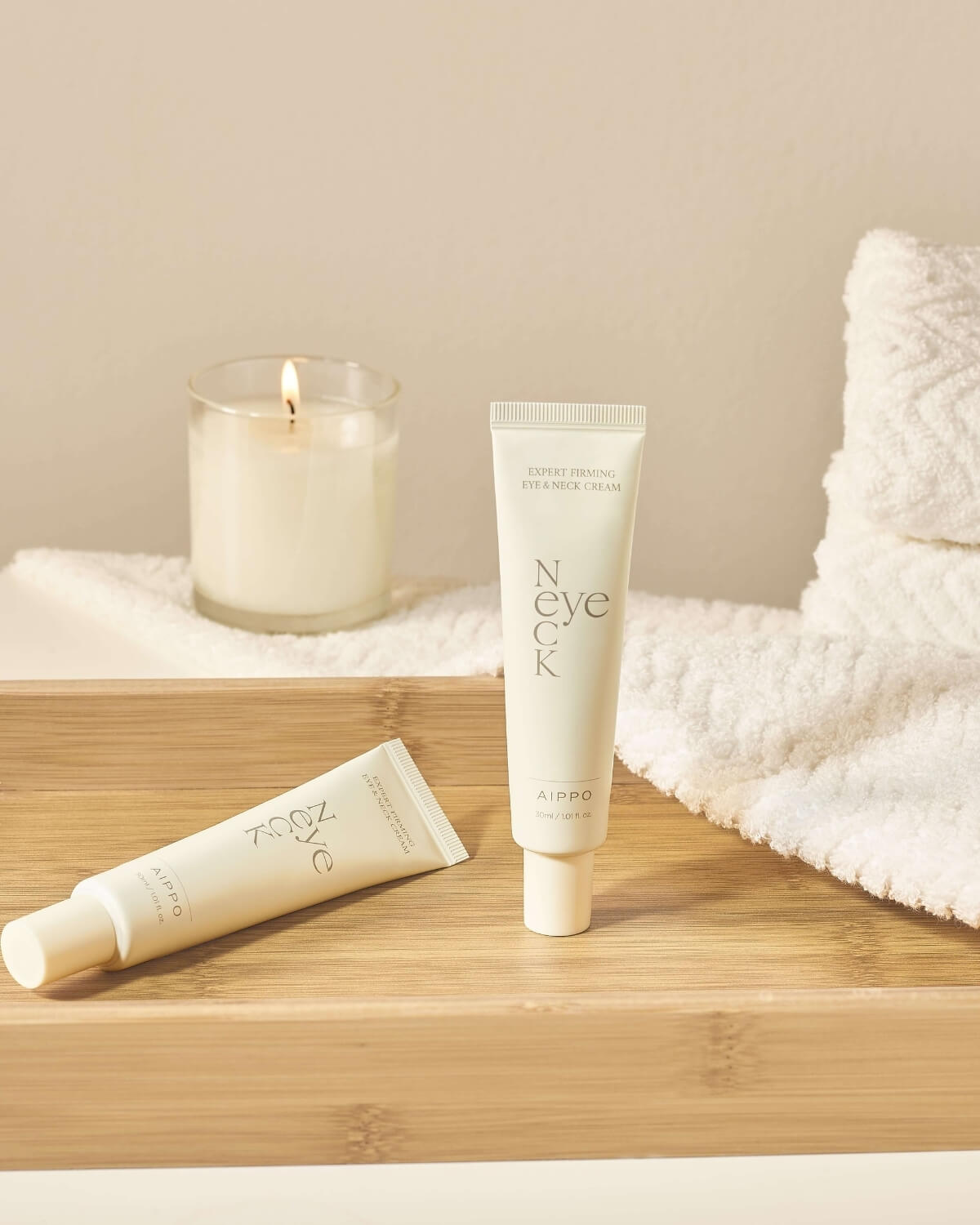 A lifestyle shot showing two tubes of the Eye & Neck Cream on a wooden tray with a stack of white rolled towels and a lit candle, suggesting a relaxing bathroom or spa setting.