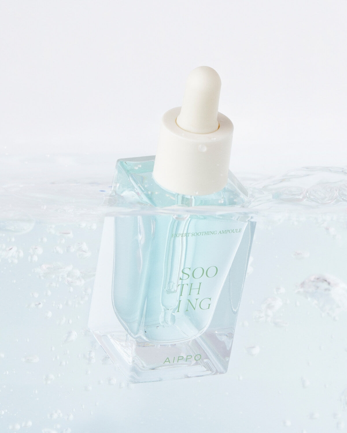 A bottle of AIPPO Expert Soothing Ampoule submerged halfway in water, creating ripples and bubbles around it. The light blue liquid inside contrasts with the white background and the surrounding clear water.