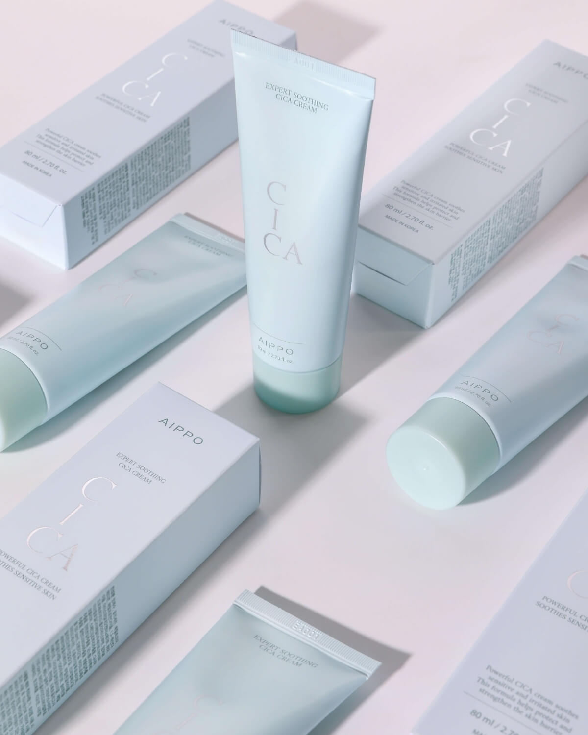 A collection of Aippo Cica Cream tubes and their packaging boxes scattered across a white surface, creating a repeating pattern. The boxes and tubes are in soft blue-green and white tones.