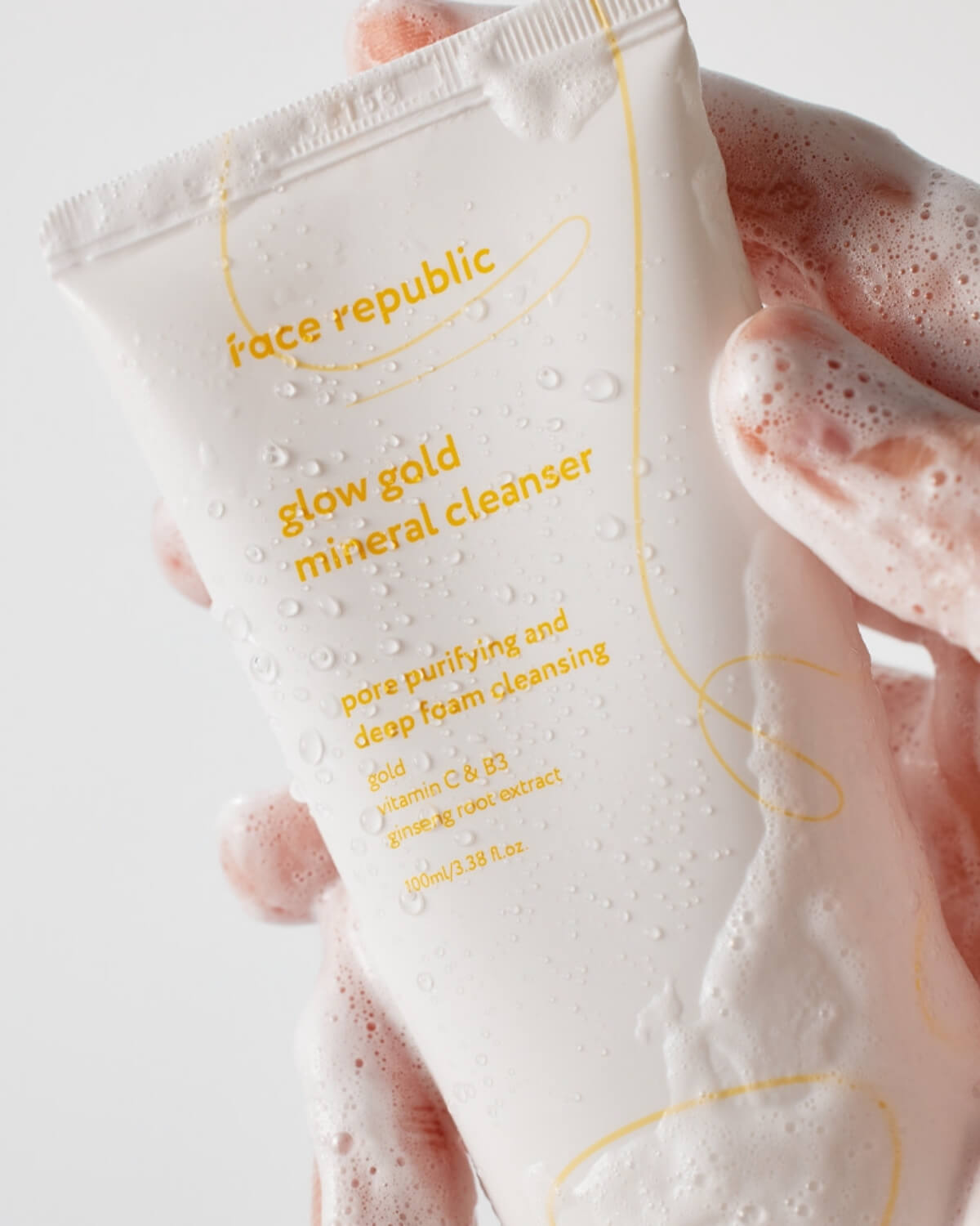 Hand holding a tube of Face Republic Glow Gold Mineral Cleanser covered in rich white foam bubbles.