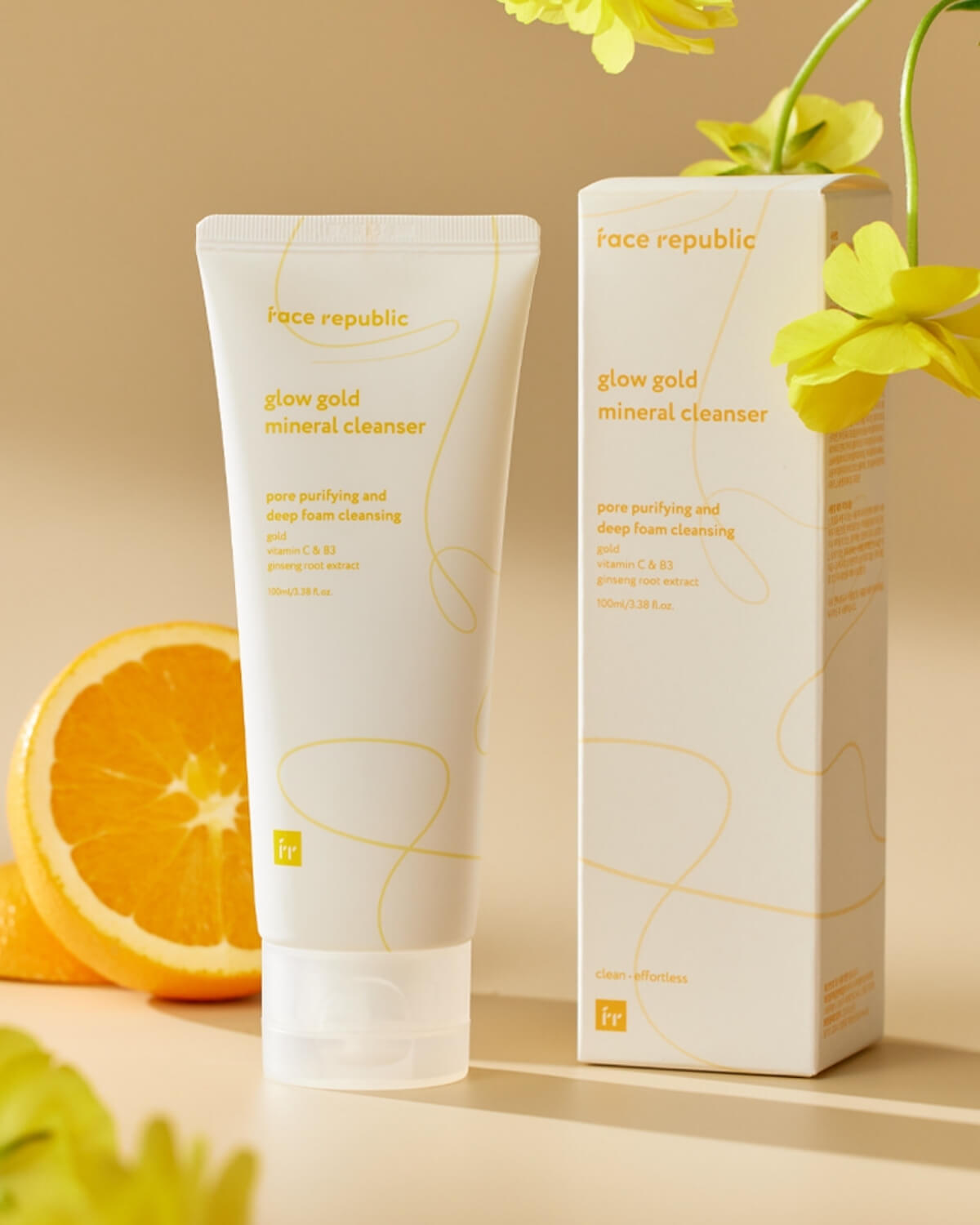 Face Republic Glow Gold Mineral Cleanser tube and packaging box next to a fresh orange slice and yellow flowers.