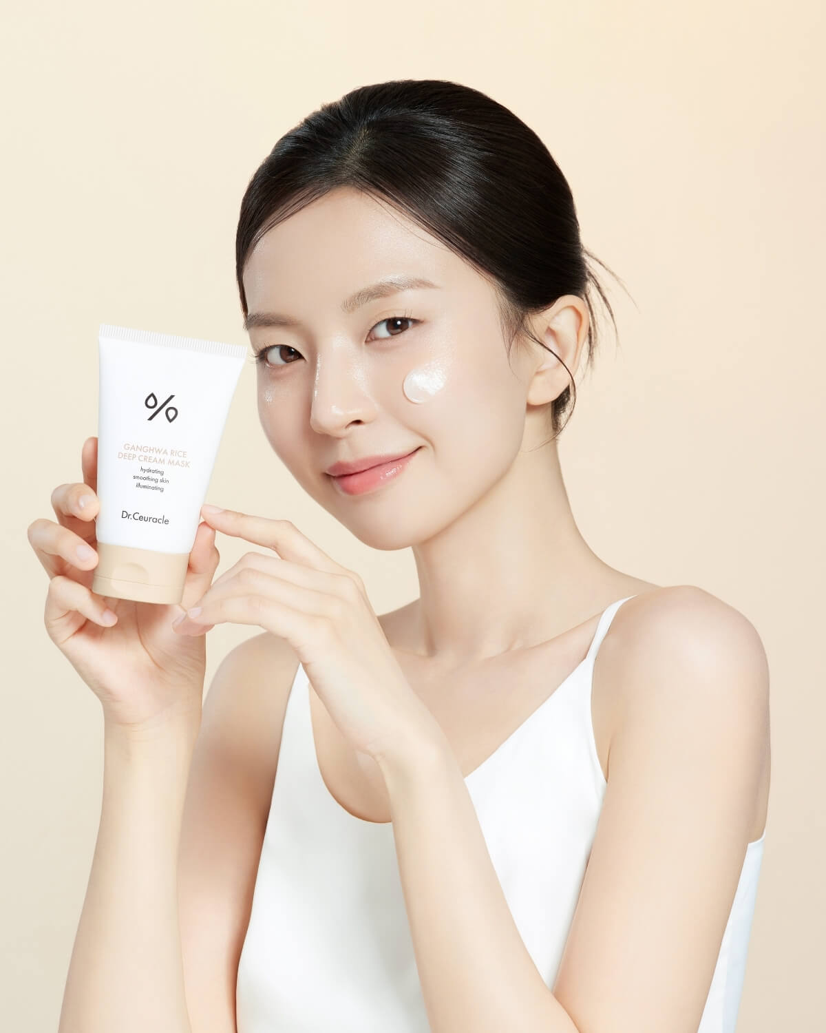 Woman with glowing skin holding Dr. Ceuracle Ganghwa Rice Deep Cream Mask with cream dabbed on cheek.