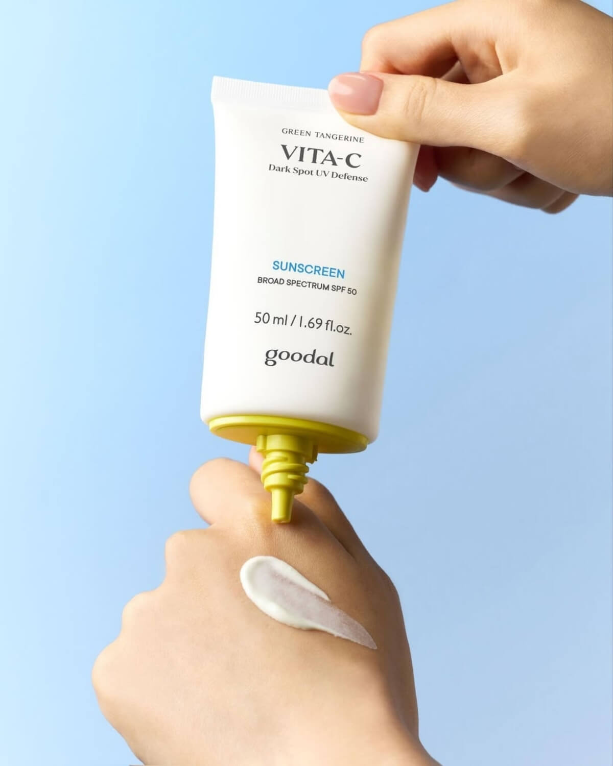A hand squeezing Goodal Vita-C sunscreen from the white and yellow tube onto the back of another hand against a blue backdrop.