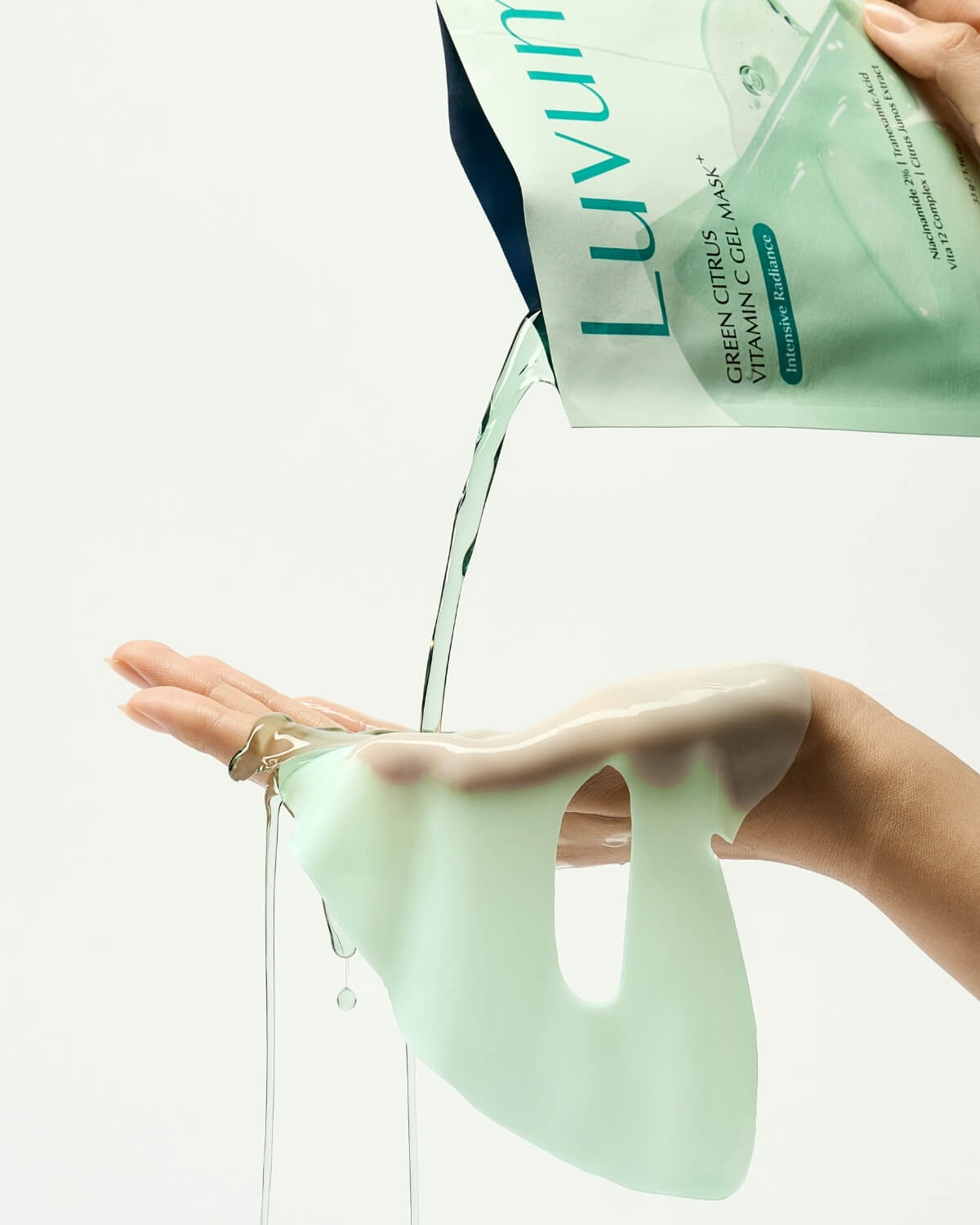 Liquid essence pouring from a packet onto a hand holding a pale green Luvum face mask sheet with eye cutouts.