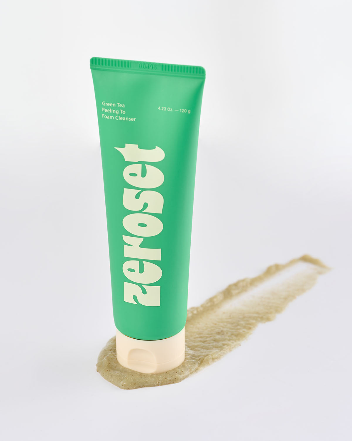 Zeroset green tea facial cleanser tube standing upright on an exfoliating gel smear on white background.