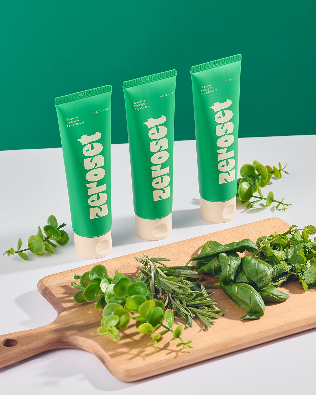 Three Zeroset green tea facial cleanser tubes lined up behind a wooden board with fresh herbs.