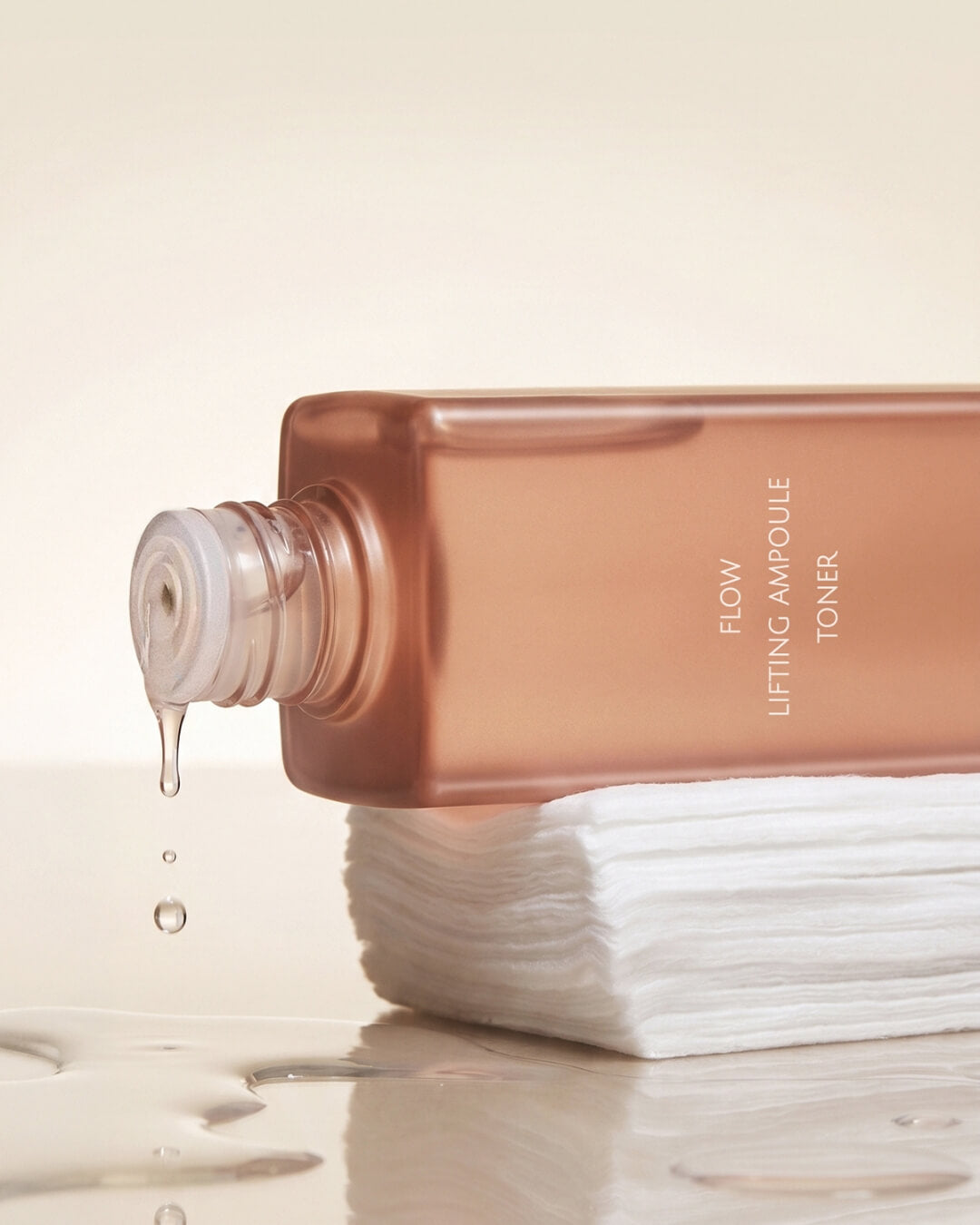 Liquid dripping from a bottle of Koy Flow Lifting Ampoule Toner onto a stack of white cotton pads.