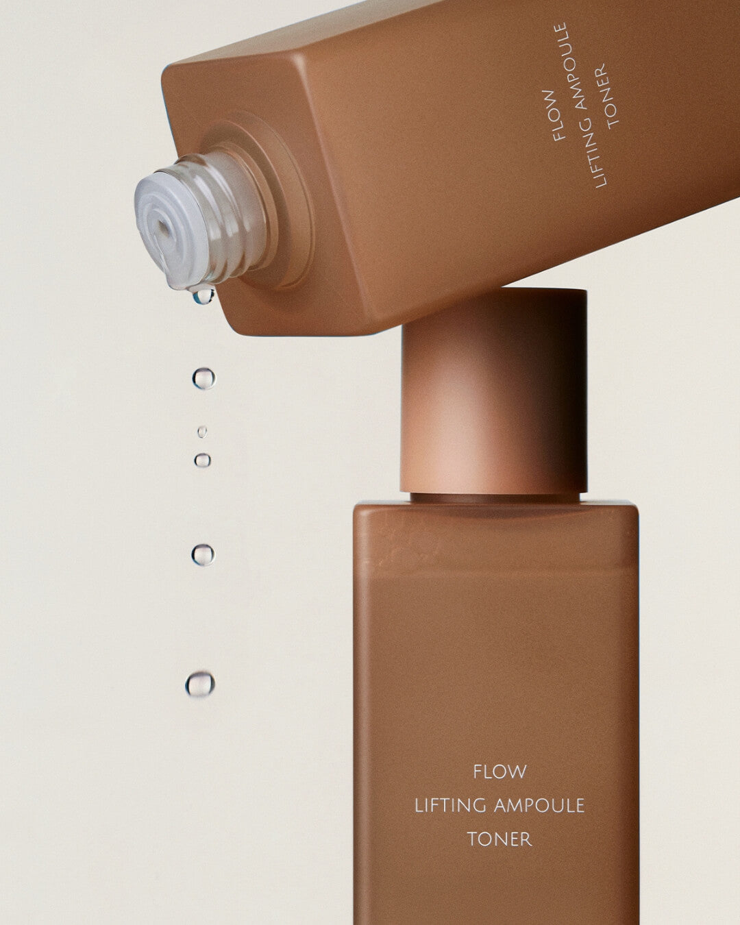 Liquid droplets falling from an inverted Koy Flow Lifting Ampoule Toner bottle onto another standing bottle.