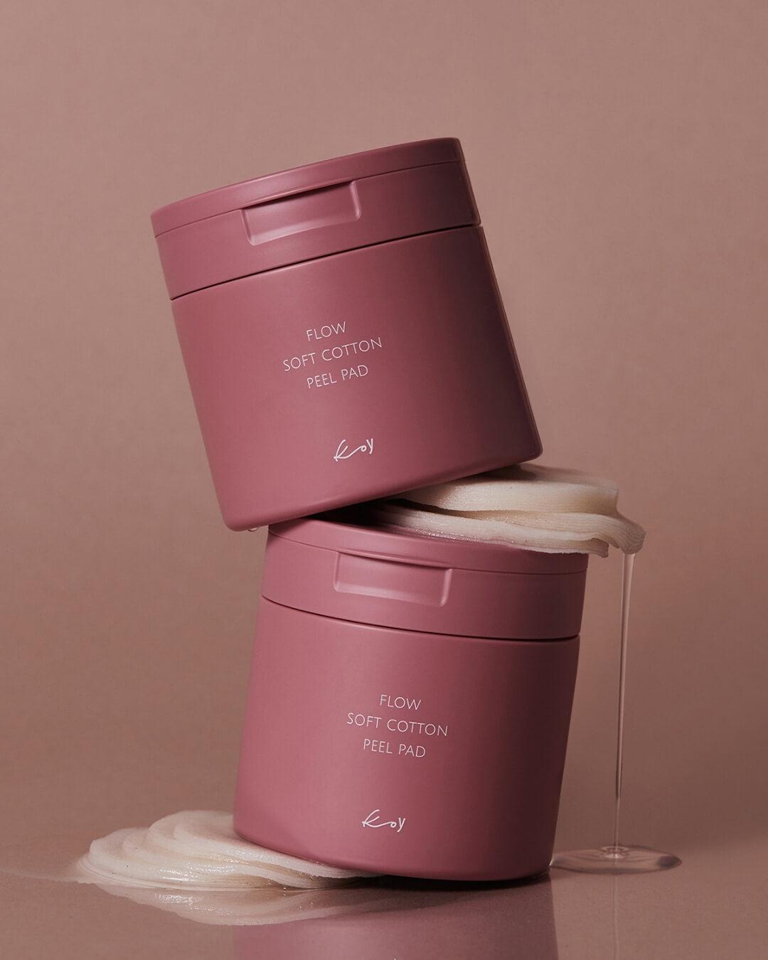 Two pink jars of Koy Flow Soft Cotton Peel Pad stacked with wet skincare pads and dripping essence.