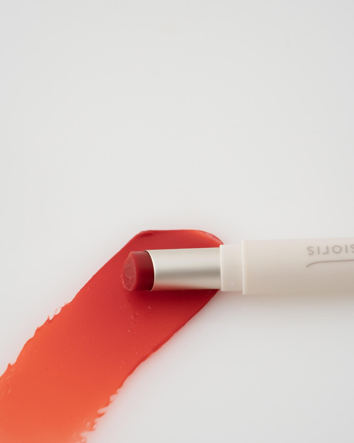 Open tube of Sioris LipSafer Color Balm in Reddish lying on top of a vibrant red-orange product swatch.