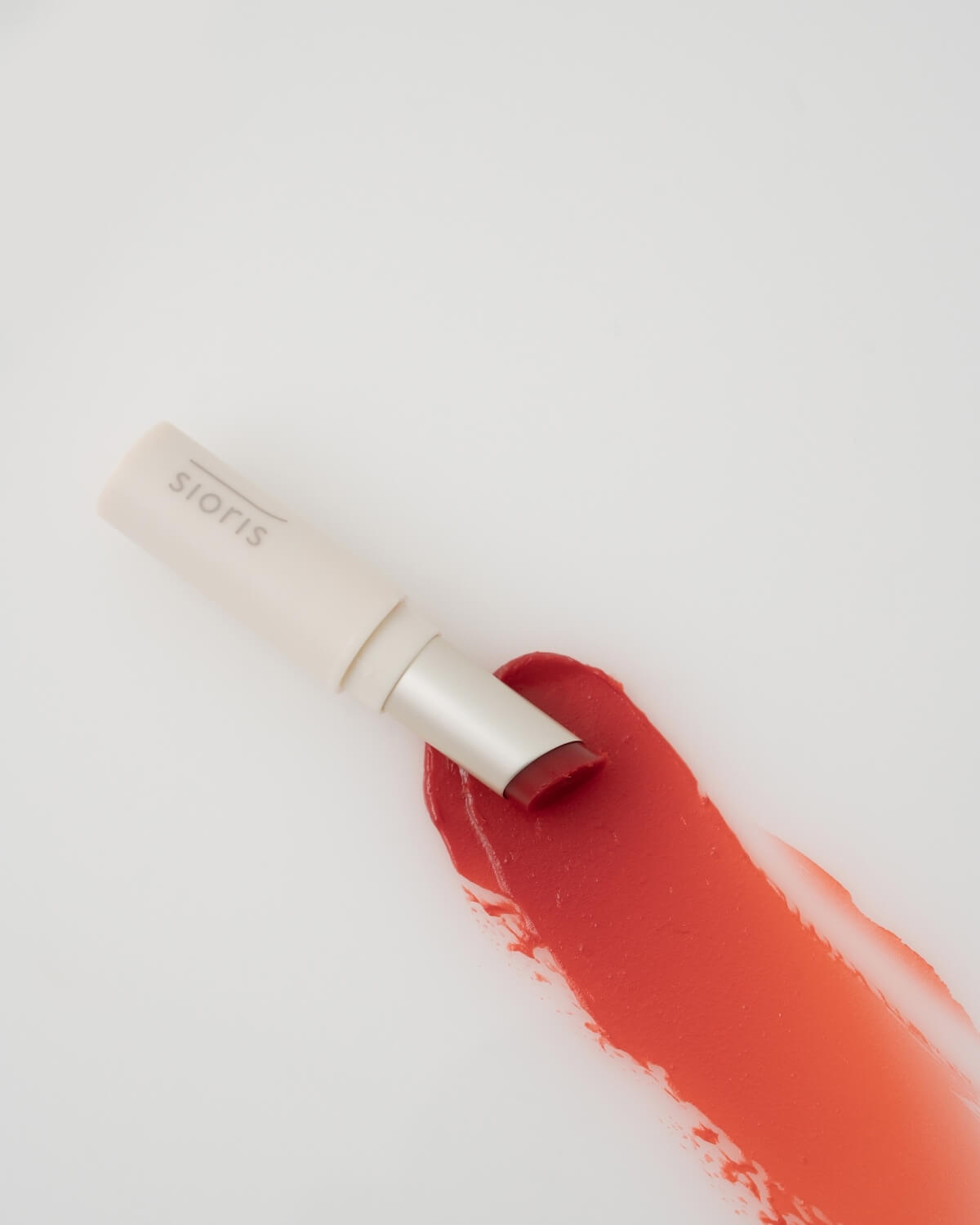 Overhead view of Sioris LipSafer Color Balm in Reddish resting diagonally over a glossy red makeup smear.