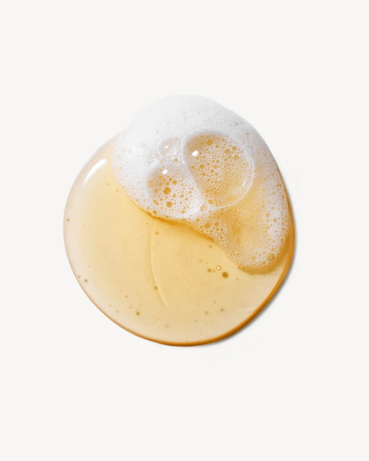 A top-down view of a clear liquid cleanser droplet with white soapy foam on a white surface.