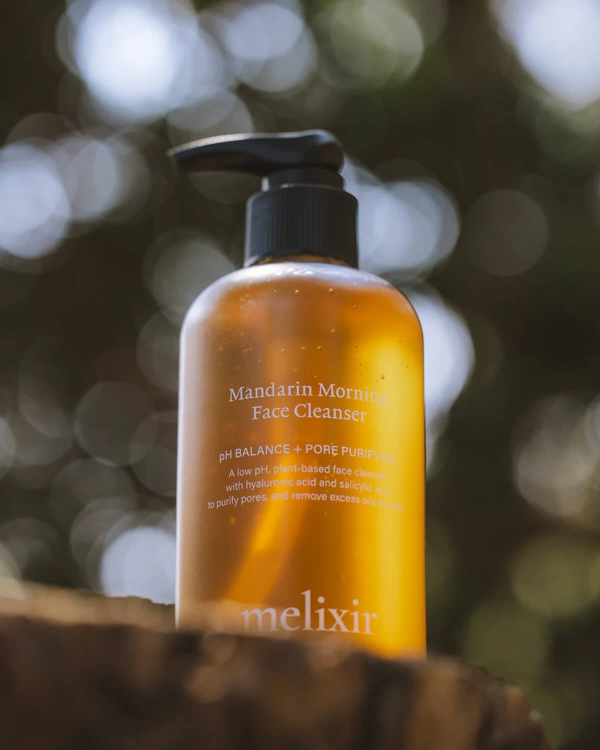 A close-up of the amber cleanser bottle outdoors with soft, blurred sunlight filtering through trees.