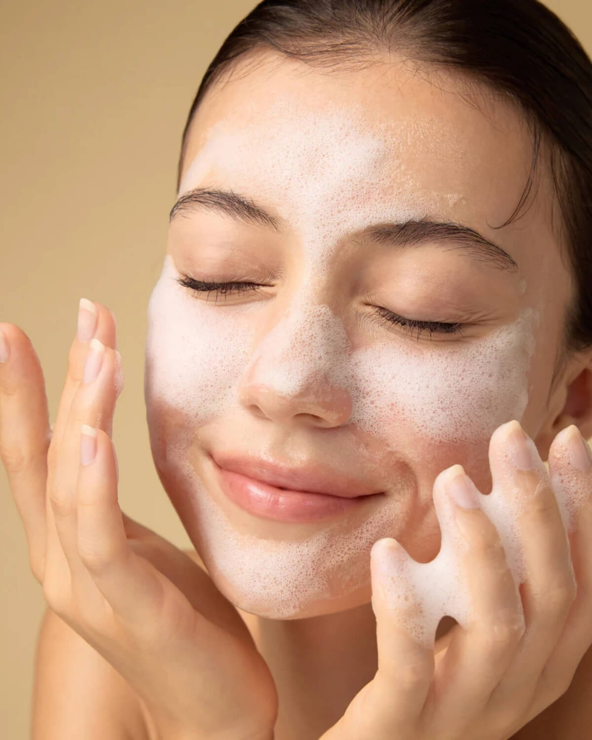 A woman smiling with her eyes closed while applying the bubbly white face cleanser to her cheeks.