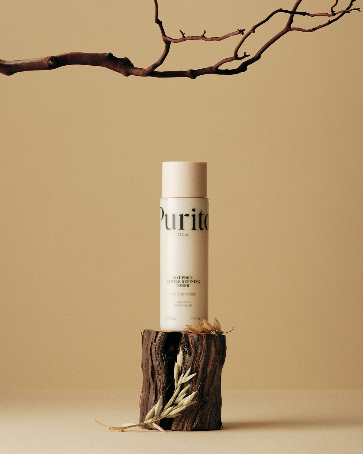 The toner bottle balanced on a piece of dark wood with an oat sprig, set under a bare branch against a beige background.