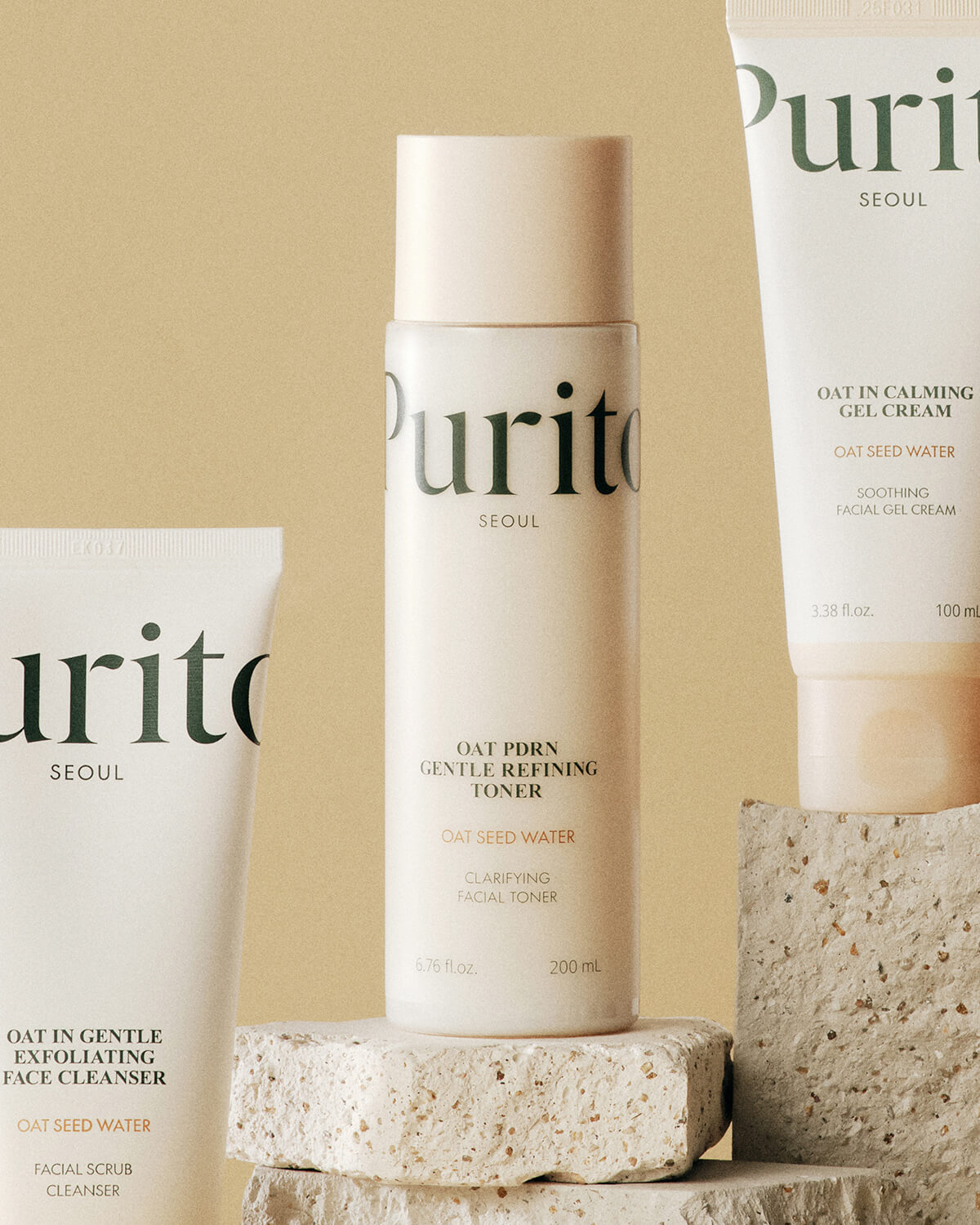 The Purito toner bottle standing on textured stone blocks, flanked by the Oat-in Gentle Exfoliating Face Cleanser and Oat-in Calming Gel Cream tubes.