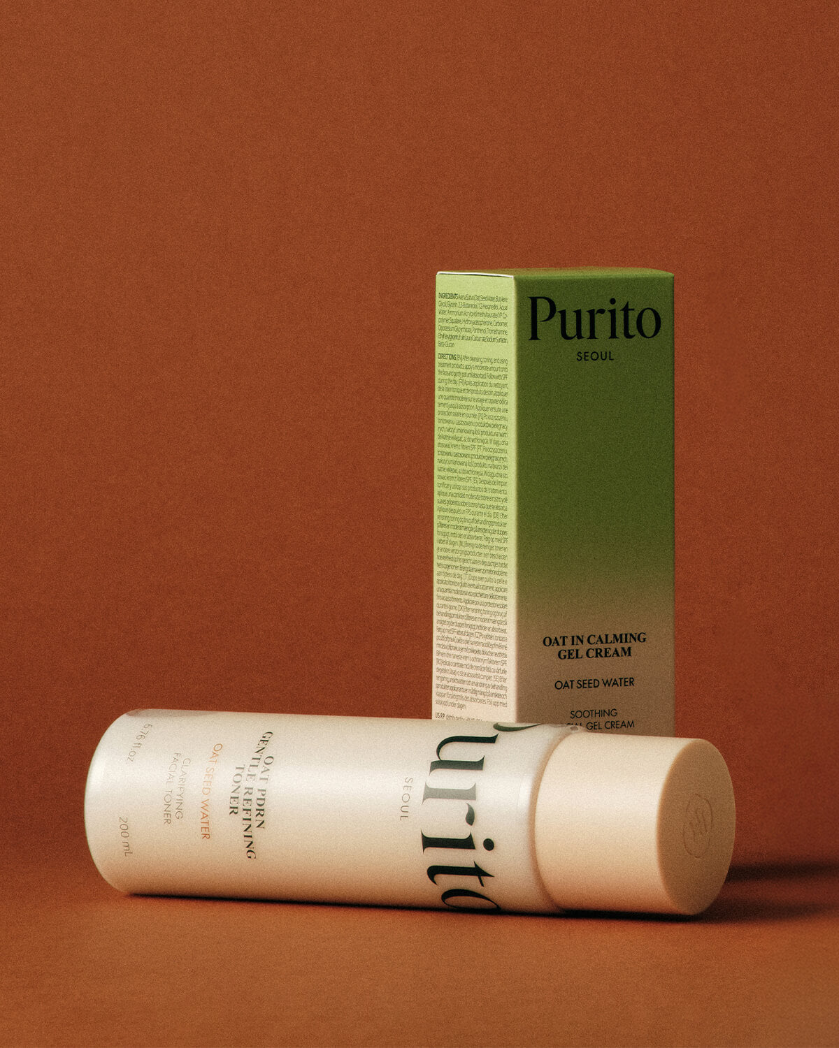 The toner bottle lying horizontally in the foreground with the Oat-in Calming Gel Cream green box standing behind it against a terracotta backdrop.