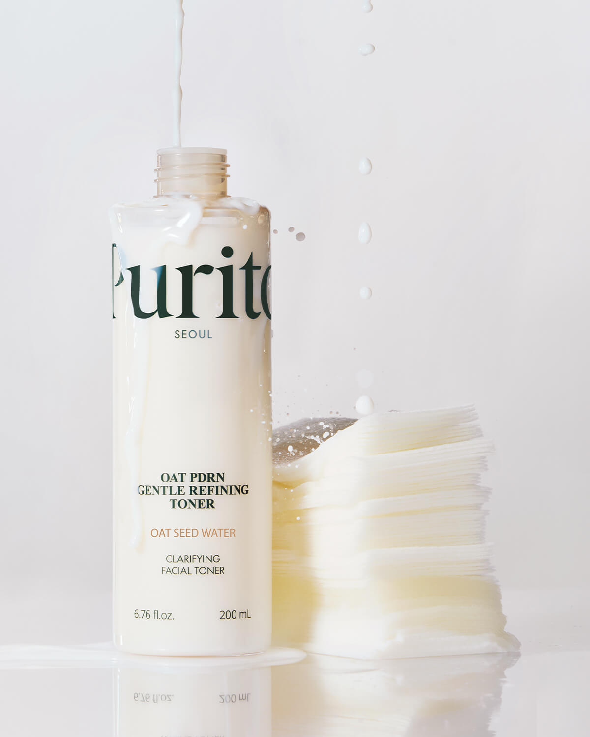 Milky toner pouring into an open bottle standing next to a stack of white cotton pads.