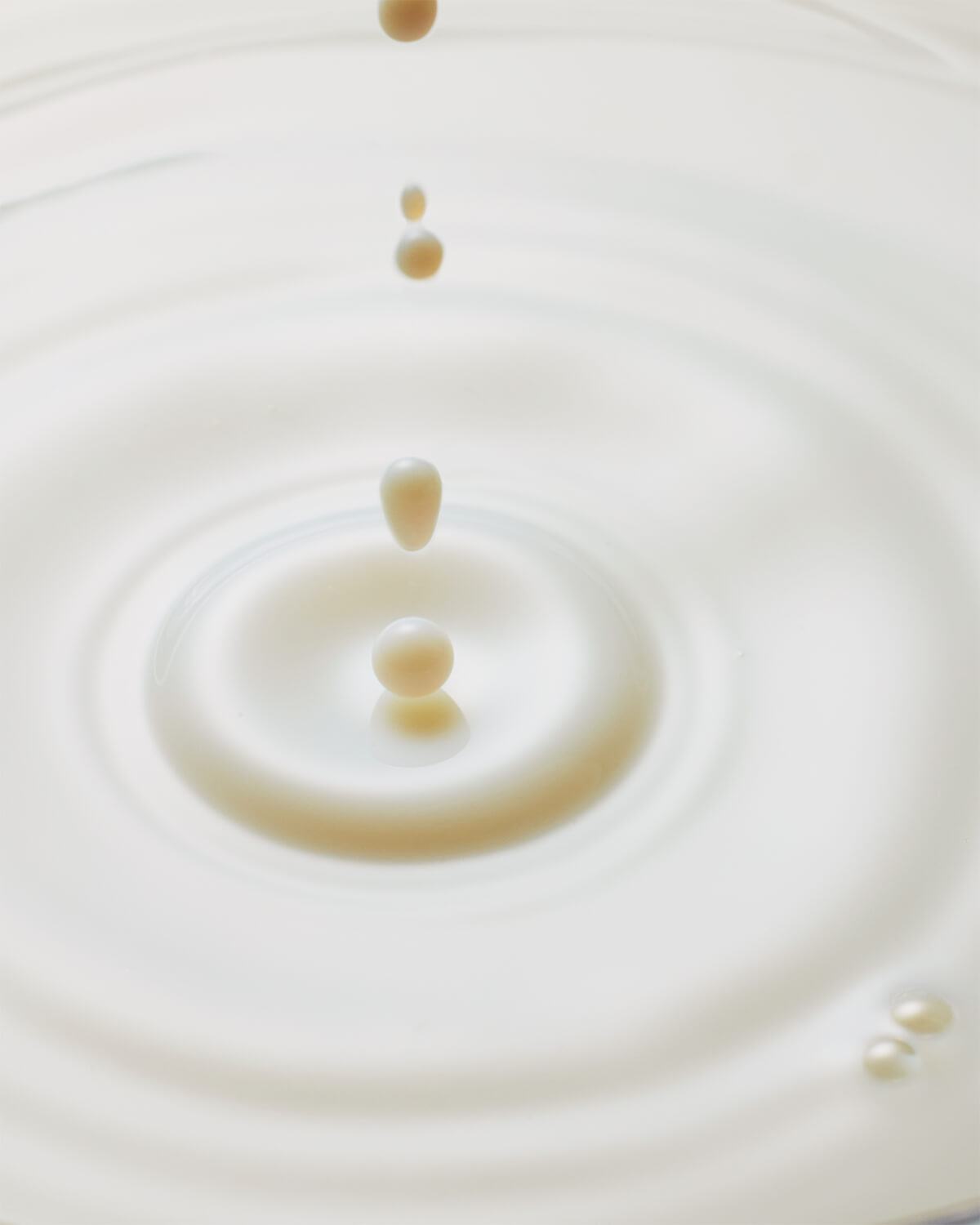 A close-up, top-down view of milky white droplets falling into a pool of liquid, creating soft concentric ripples.