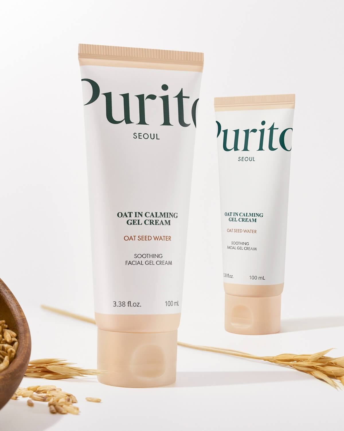 Two Purito Oat-in Calming Gel Cream tubes displayed next to a wooden bowl of oats and wheat stalks.
