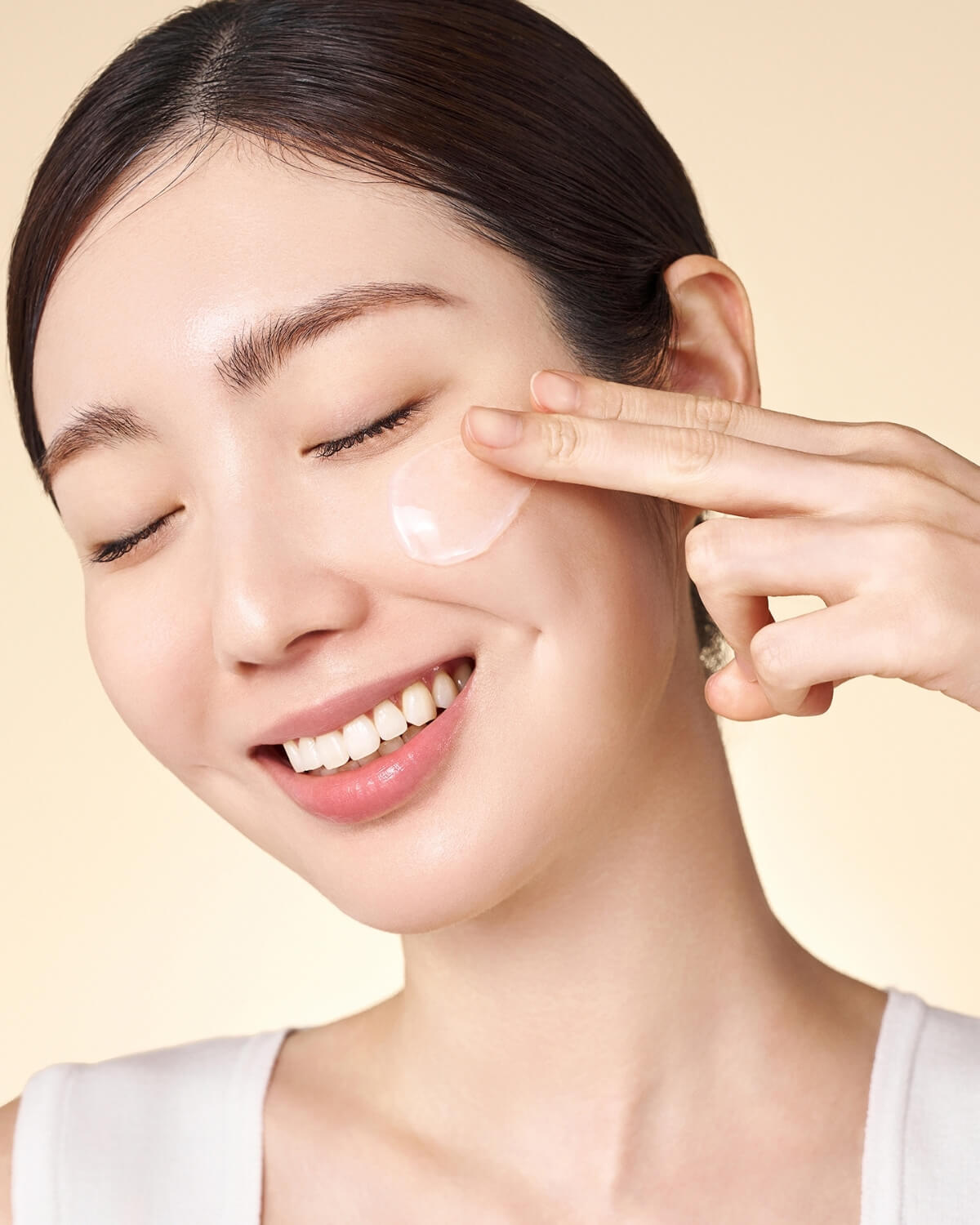 Close-up of a smiling woman gently applying a drop of clear gel cream to her cheek with two fingers.