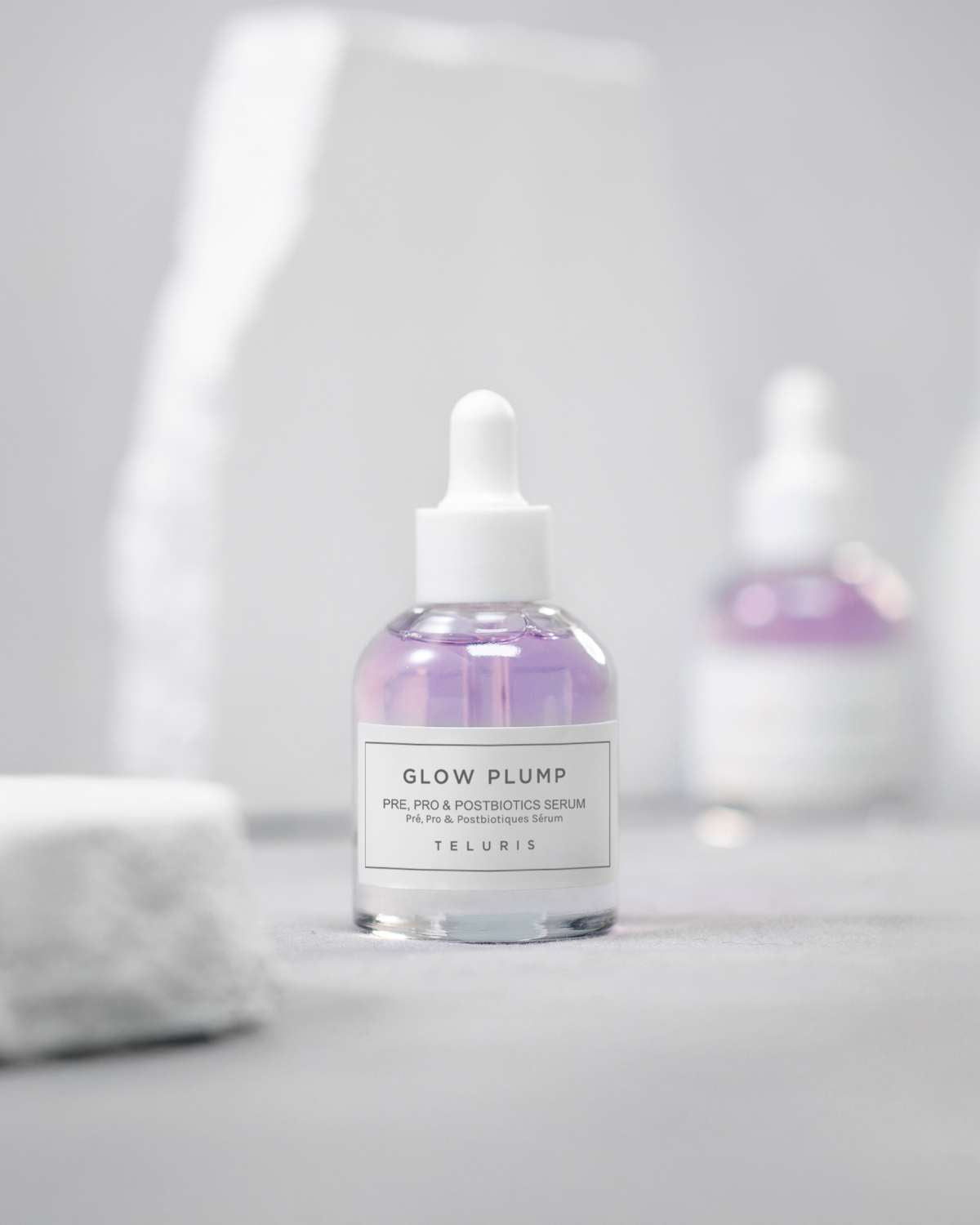 Pink Teluris Glow Plump Serum bottle displayed on a white stone pedestal with a grey background.