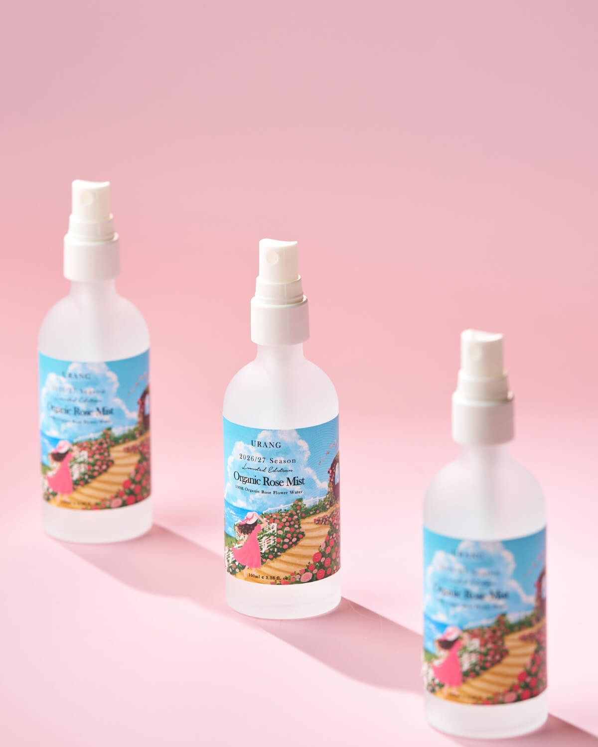 Three URANG Organic Rose Mist spray bottles in a row on a soft pink background with dramatic shadows.