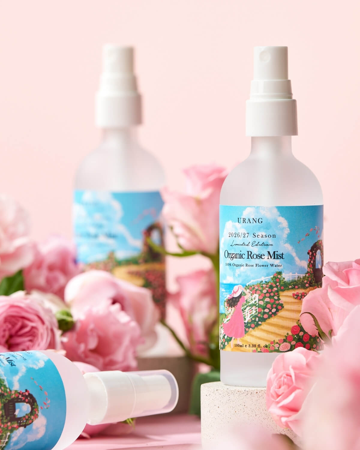 URANG Organic Rose Mist bottles arranged on pedestals with fresh pink roses on a pink studio setup.