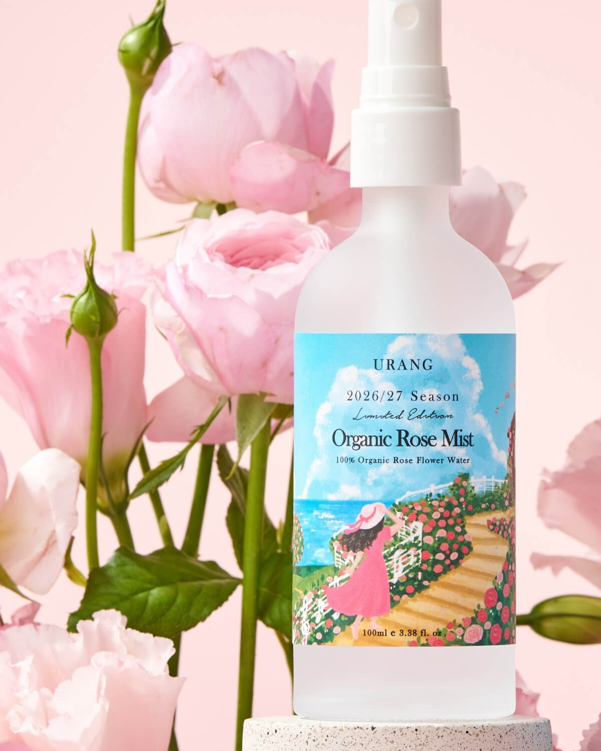 Close-up of URANG Organic Rose Mist bottle standing on a stone base among pink roses and green stems.