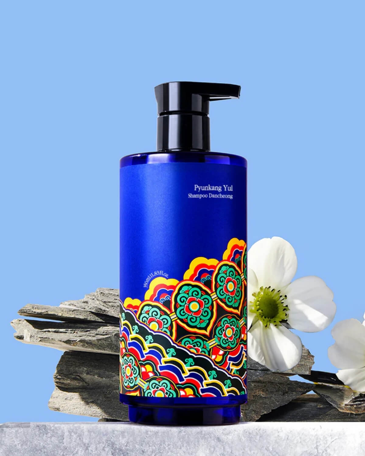 Pyunkang Yul Shampoo Dancheong bottle displayed on gray slate rocks with white flowers and a light blue background for a natural, editorial look.