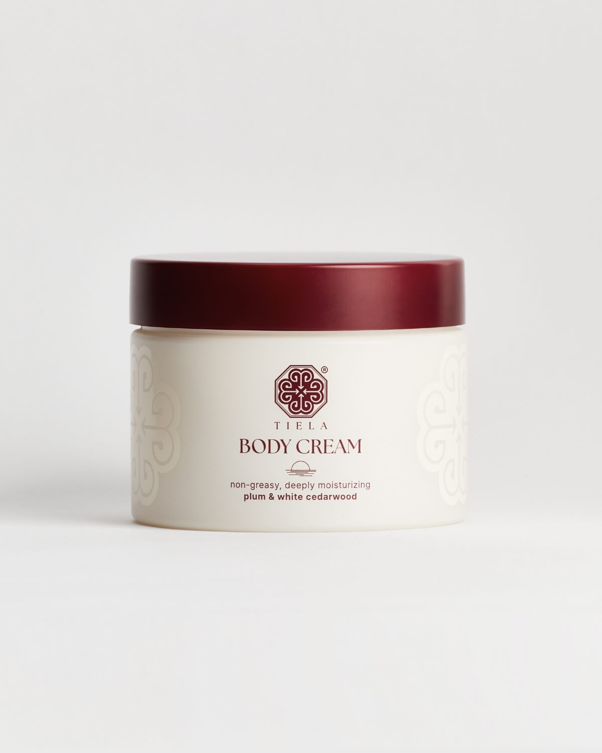 Front view of a round Tiela Body Cream jar with a deep red lid and white container, labeled "non-greasy, deeply moisturizing plum & white cedarwood."