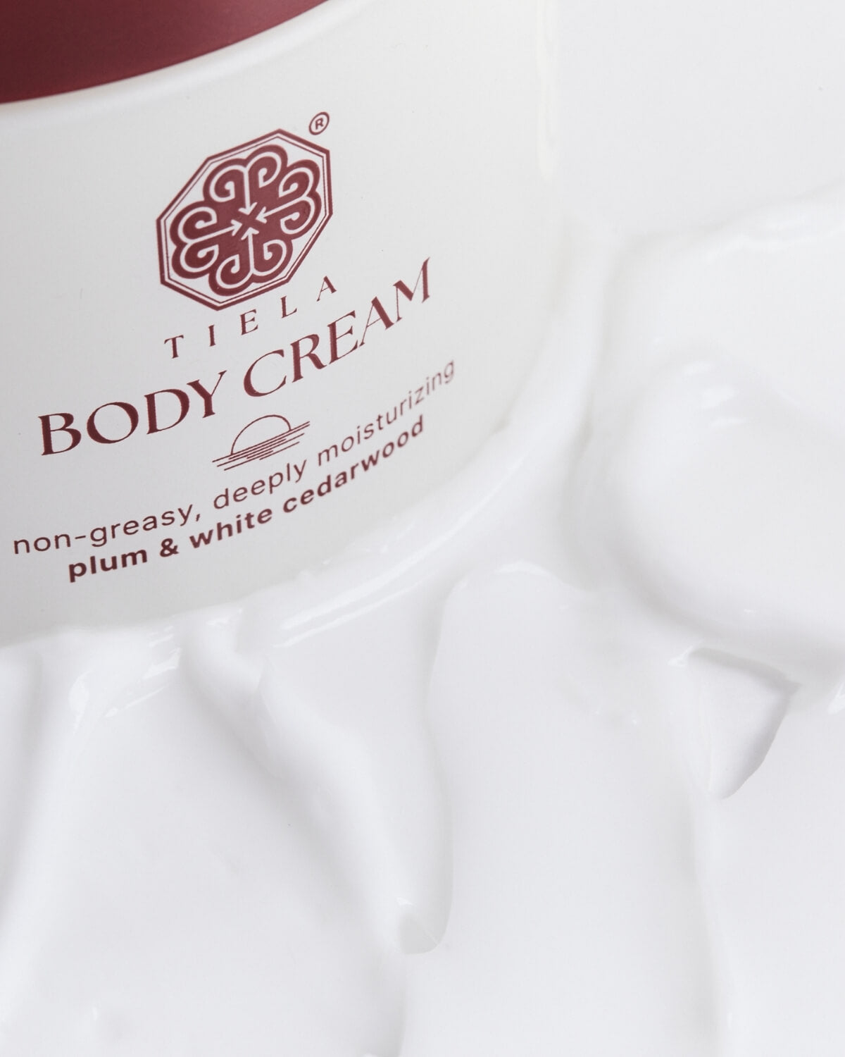 Extreme close-up of the Tiela Body Cream label ("Tiela Body Cream non-greasy, deeply moisturizing plum & white cedarwood") surrounded by the thick white cream.