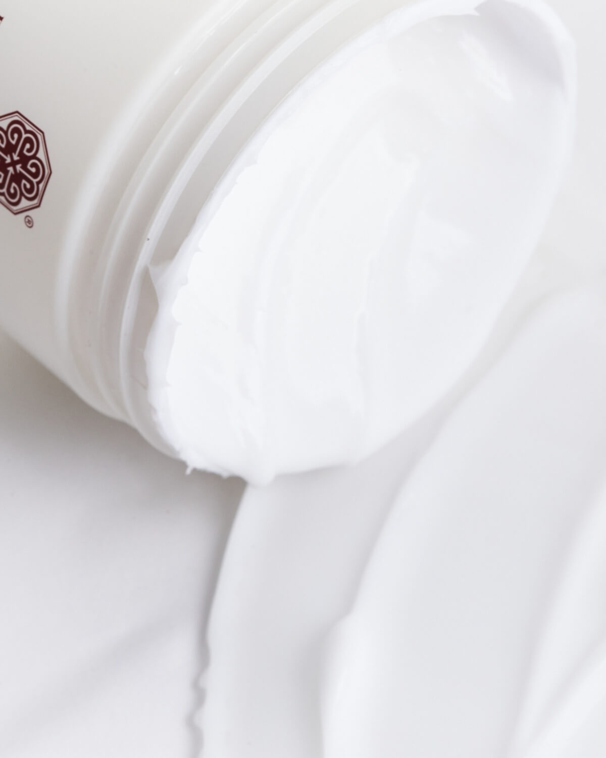 Close-up of the Tiela Body Cream jar tilted, showing a generous amount of the thick, white cream spilling out onto a smooth white surface.