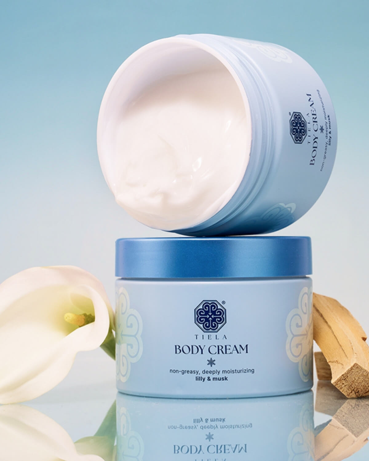 Two light blue Tiela Body Cream jars stacked, with the top jar open to show the white cream. The background includes a white calla lily and a piece of light-colored wood.