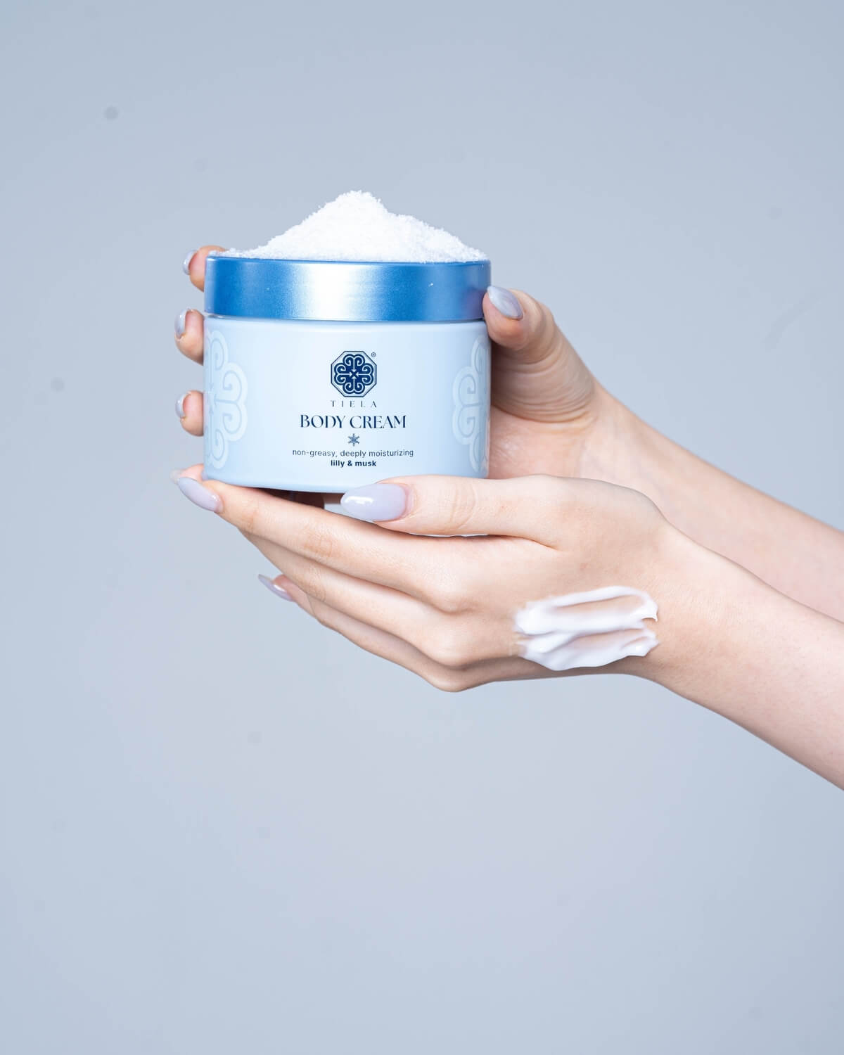 A pair of hands holding a light blue Tiela Body Cream jar, which is overflowing with white crystalline material. A swipe of cream is on the back of one hand.
