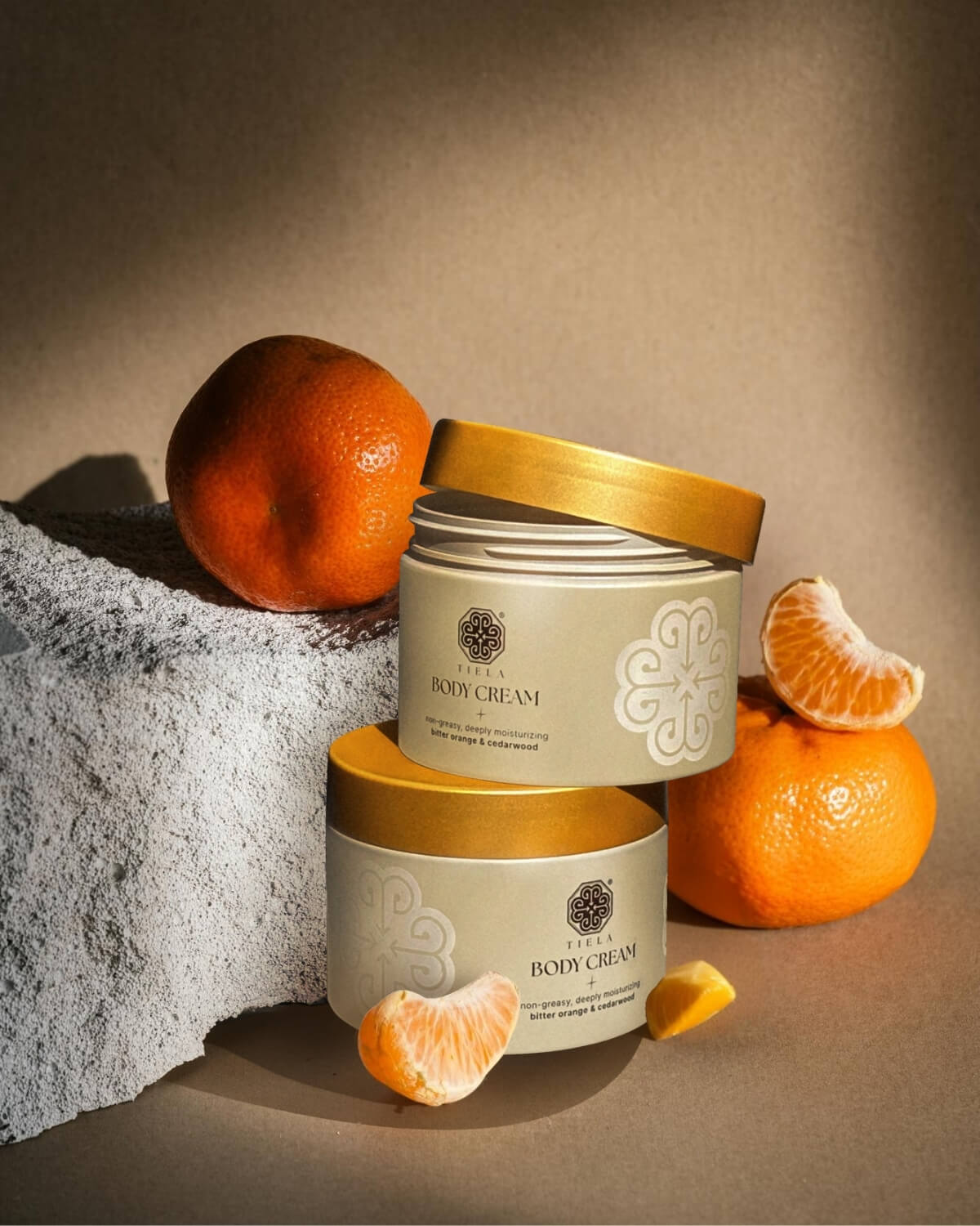 Two beige and gold Tiela Body Cream jars stacked on a rough white stone, surrounded by whole and segmented mandarin oranges.