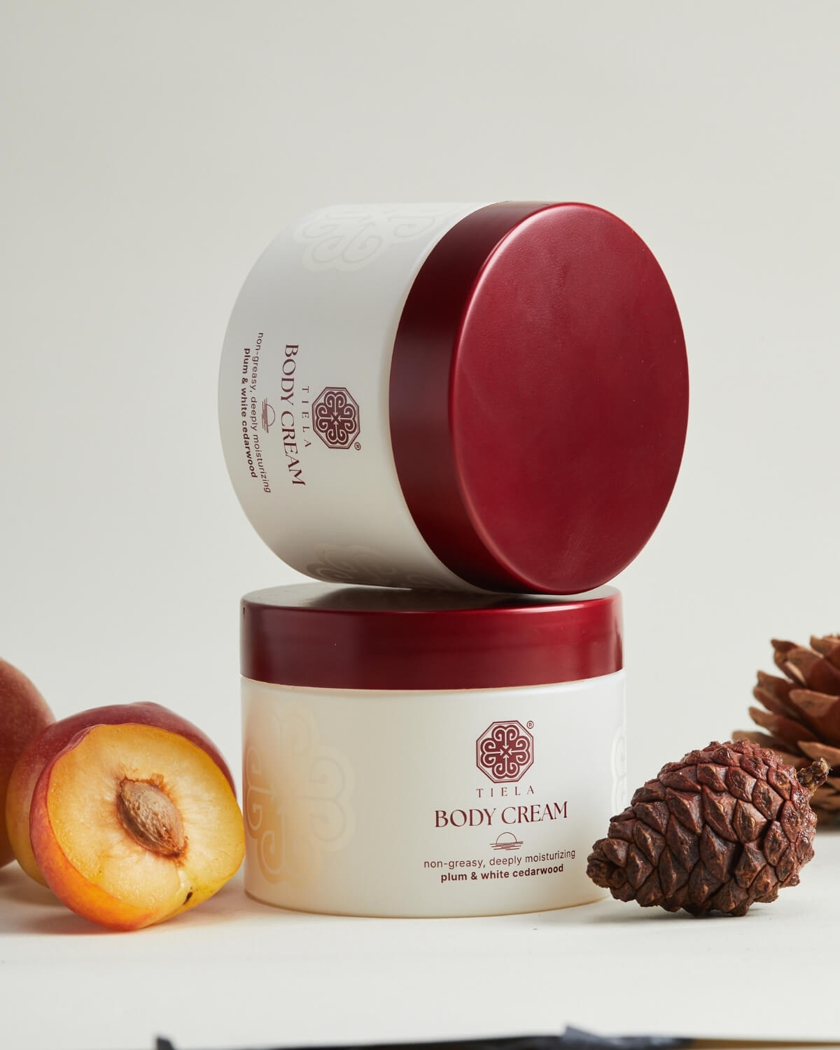 Two Tiela Body Cream jars stacked vertically, with the lids closed, surrounded by a halved plum and pine cones.