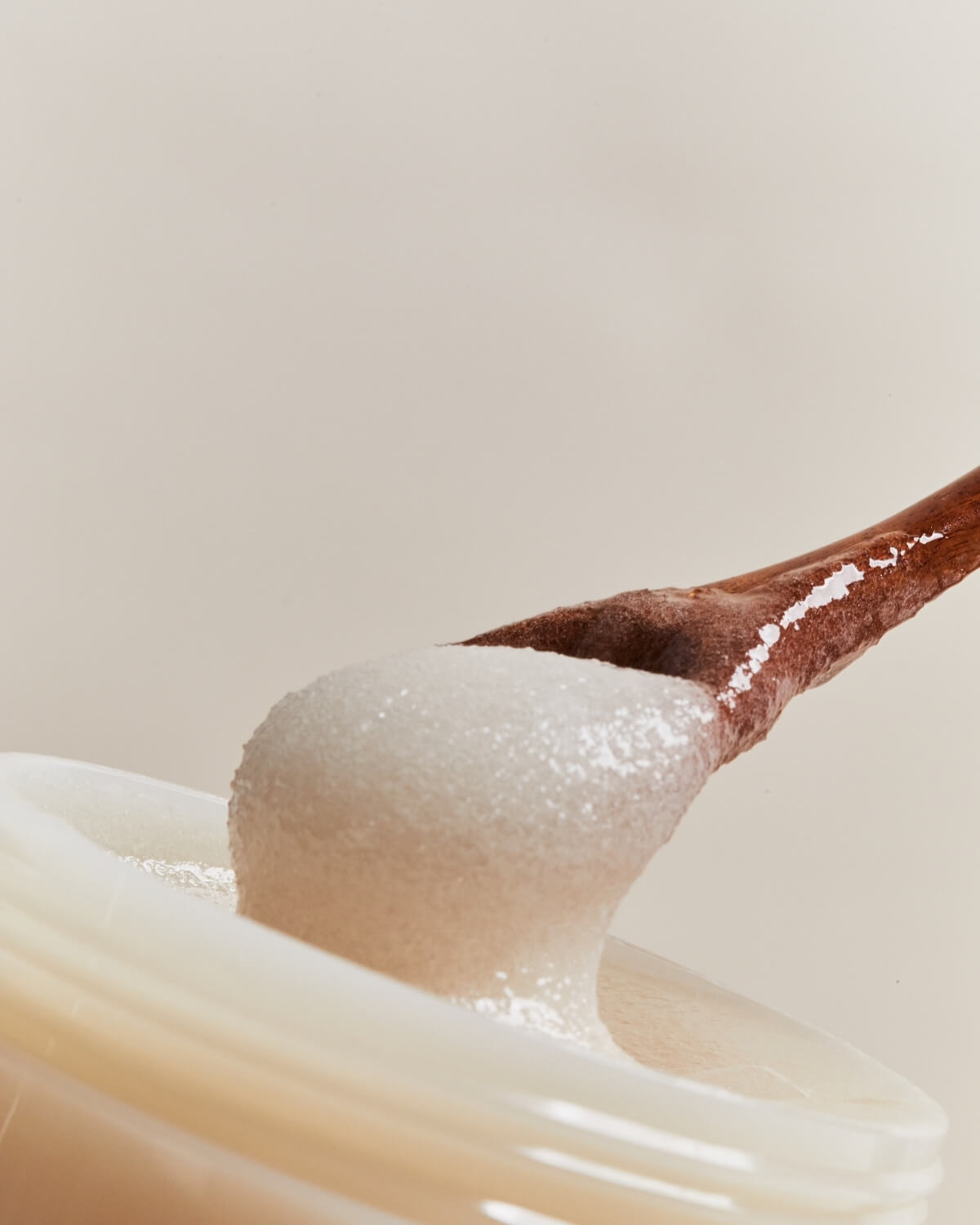 Extreme close-up of a wooden spoon scooping out the white, grainy body scrub product from the jar.