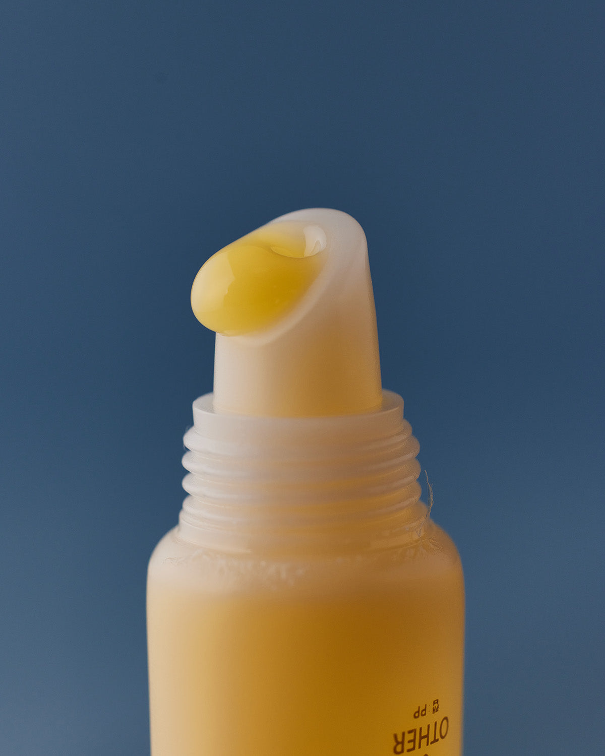 An extreme close-up of a yellowish product dispenser or pump, showing a drop of the thick, oily, yellow-gold Jojoba Butter Balm resting on the nozzle. The background is a solid, deep blue.
